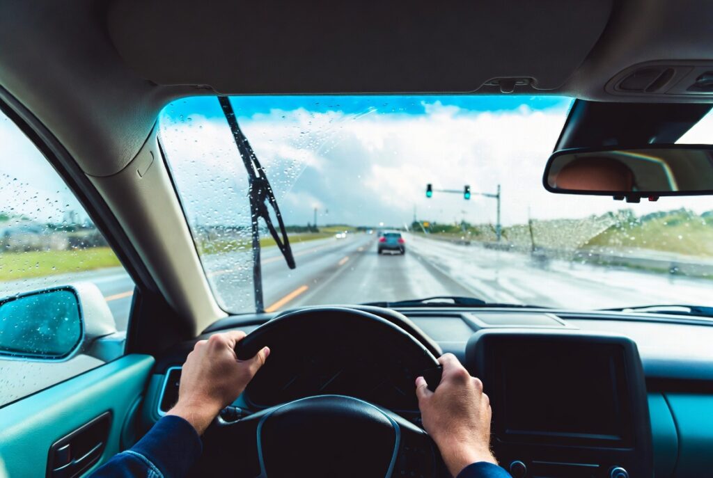 First-person view from inside a car during light rain, showing steady hands on the steering wheel and safe defensive driving on a wet Texas highway with good visibility.